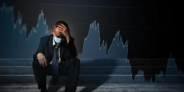 Man in suit and face mask, with a worried expression, sitting in front of a declining graph.